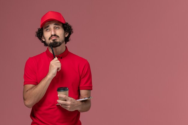 Vista frontal repartidor de mensajería masculino en camisa roja y capa sosteniendo una taza de café marrón y un bloc de notas con bolígrafo pensando en el trabajo de empleado de entrega de servicio de pared rosa claro