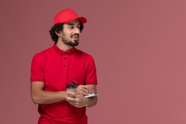 Vista frontal repartidor de mensajería masculino en camisa roja y capa sosteniendo una taza de café marrón y un bloc de notas en la pared rosa claro empleado de servicio de entrega