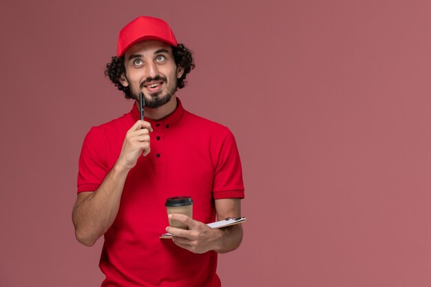 Vista frontal repartidor de mensajería masculino en camisa roja y capa sosteniendo una taza de café marrón y un bloc de notas pensando en el empleado de entrega uniforme de servicio de pared rosa