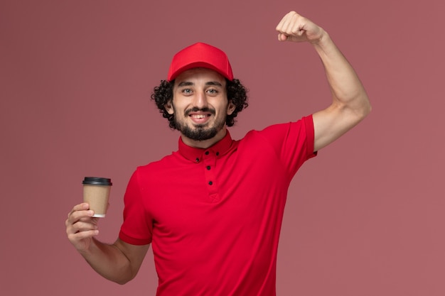 Vista frontal repartidor de mensajería masculino en camisa roja y capa sosteniendo una taza de café marrón y flexionando en el empleado de entrega uniforme de servicio de pared rosa claro