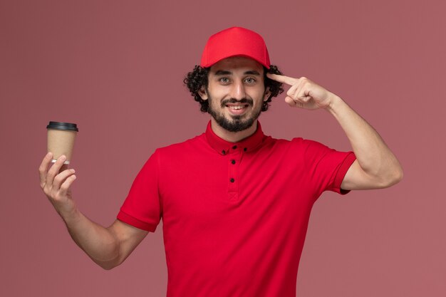 Vista frontal repartidor de mensajería masculino en camisa roja y capa sosteniendo una taza de café marrón en la pared de color rosa claro, servicio de entrega uniforme, empleado, trabajo masculino