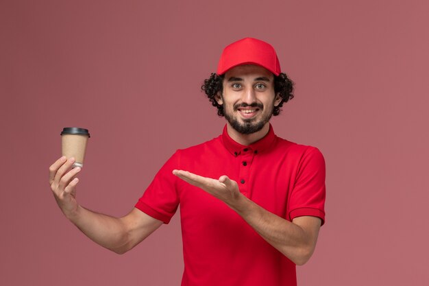 Vista frontal repartidor de mensajería masculino en camisa roja y capa sosteniendo una taza de café marrón en la pared de color rosa claro trabajador empleado de entrega uniforme de servicio