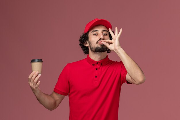 Vista frontal repartidor de mensajería masculino en camisa roja y capa sosteniendo una taza de café marrón en la pared rosa claro empleado de entrega uniforme de servicio