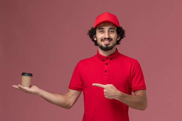 Vista frontal repartidor de mensajería masculino en camisa roja y capa sosteniendo una taza de café marrón en la pared rosa servicio uniforme de entrega empleado trabajo masculino