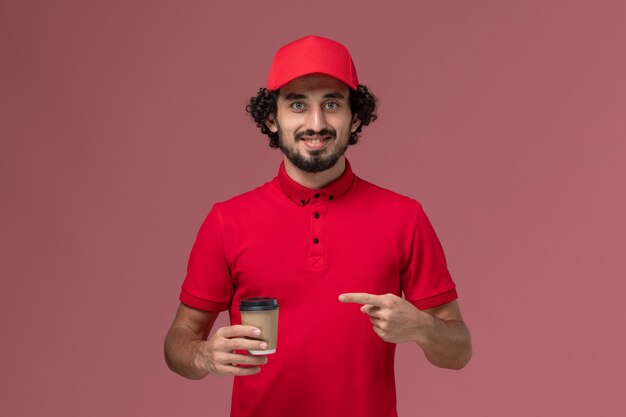 Vista frontal repartidor de mensajería masculino en camisa roja y capa sosteniendo una taza de café marrón en la pared rosa servicio uniforme de entrega trabajo empleado trabajo masculino