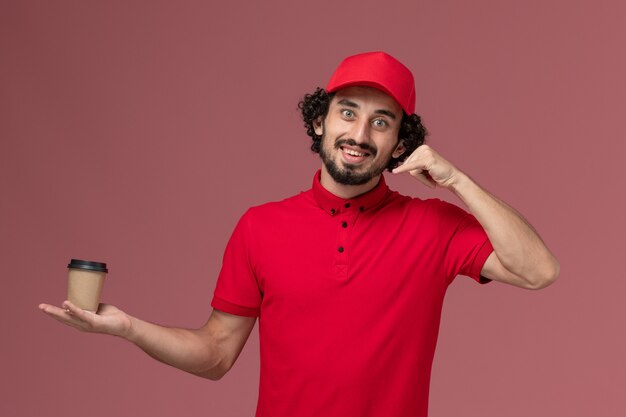 Vista frontal repartidor de mensajería masculino en camisa roja y capa sosteniendo una taza de café marrón en la pared rosa servicio uniforme trabajador empleado de entrega