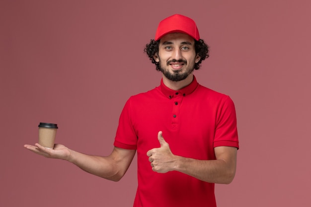 Vista frontal repartidor de mensajería masculino en camisa roja y capa sosteniendo una taza de café marrón en la pared rosa servicio uniforme de trabajo de los empleados