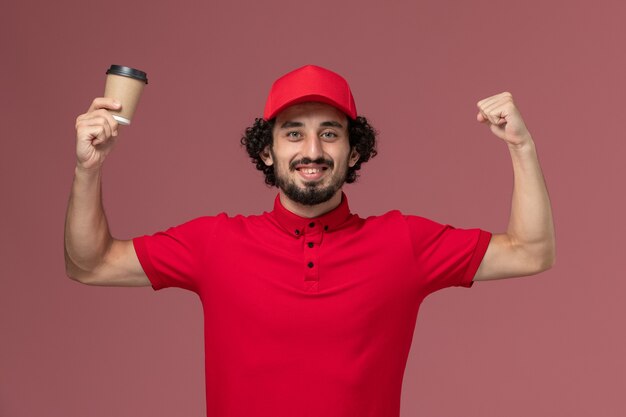 Vista frontal repartidor de mensajería masculino en camisa roja y capa sosteniendo una taza de café marrón y sonriendo en el empleado de entrega uniforme de servicio de pared rosa claro