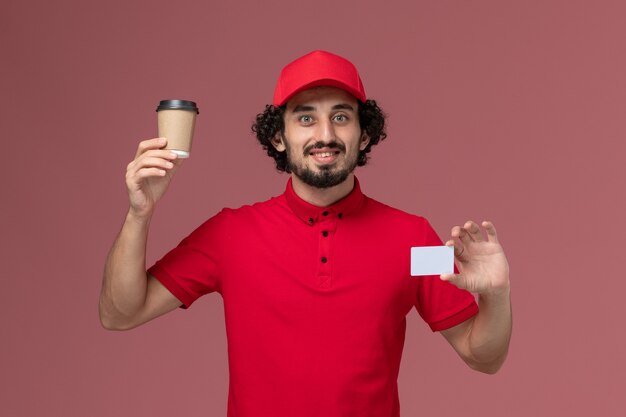 Vista frontal repartidor de mensajería masculino en camisa roja y capa sosteniendo una taza de café marrón con tarjeta en la pared rosa claro trabajo de empleado de entrega uniforme