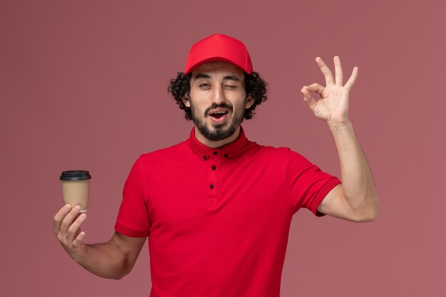 Vista frontal repartidor de mensajería masculino en camisa roja y capa sosteniendo una taza de café marrón en el trabajo de empleado de entrega uniforme de servicio de pared rosa claro