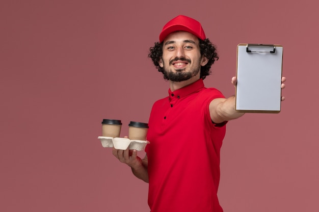 Vista frontal repartidor de mensajería masculino en camisa roja y capa sosteniendo tazas de café de entrega marrón y bloc de notas en la pared rosa claro empleado de repartidor de servicio