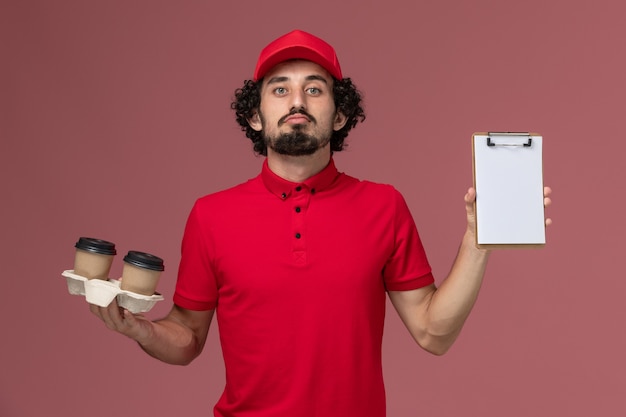 Vista frontal repartidor de mensajería masculino en camisa roja y capa sosteniendo tazas de café de entrega marrón y bloc de notas en la pared rosa claro empleado de servicio de entrega