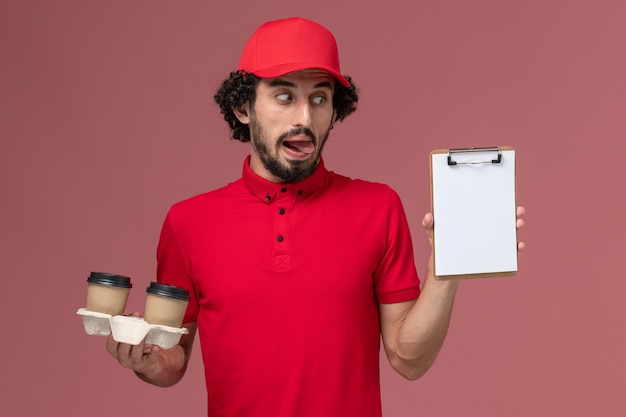 Vista frontal repartidor de mensajería masculino en camisa roja y capa sosteniendo tazas de café de entrega marrón y bloc de notas en la pared rosa claro trabajo empleado de entrega de servicio masculino