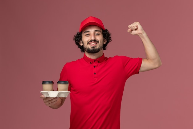 Vista frontal repartidor de mensajería masculino en camisa roja y capa sosteniendo tazas de café de entrega marrón en empleado de entrega de servicio de trabajador de pared rosa