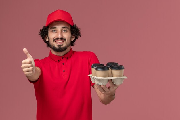 Foto gratuita vista frontal repartidor de mensajería masculino en camisa roja y capa sosteniendo tazas de café de entrega marrón en la empresa de empleados de entrega de servicios de pared rosa claro