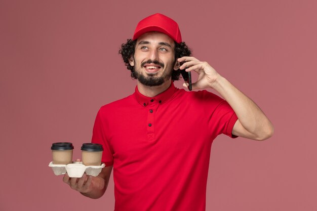 Vista frontal repartidor de mensajería masculino en camisa roja y capa sosteniendo tazas de café de entrega marrón y hablando por teléfono en la pared rosa claro trabajador empleado de servicio de entrega