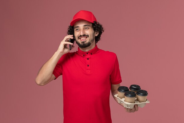 Vista frontal repartidor de mensajería masculino con camisa roja y capa sosteniendo tazas de café de entrega marrón junto con el teléfono hablando por el empleado de trabajo de entrega de servicio de pared rosa