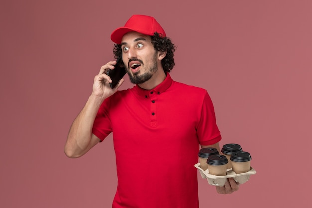 Vista frontal repartidor de mensajería masculino en camisa roja y capa sosteniendo tazas de café de entrega marrón junto con el teléfono en el trabajo de empleado de entrega de servicio de pared rosa