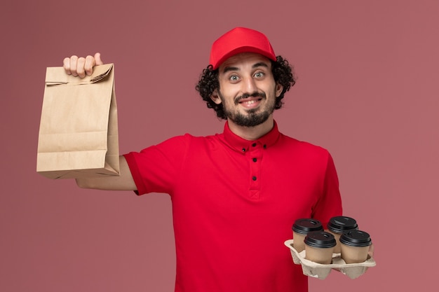Vista frontal repartidor de mensajería masculino en camisa roja y capa sosteniendo tazas de café de entrega marrón con paquete de comida en la pared rosada empleado de servicio de entrega masculino
