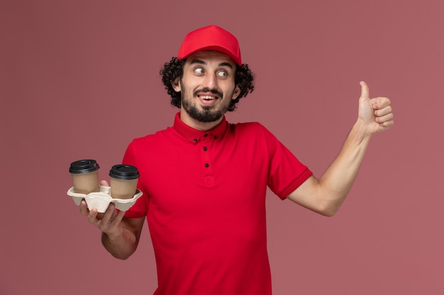 Vista frontal repartidor de mensajería masculino con camisa roja y capa sosteniendo tazas de café de entrega marrón en la pared rosa claro empleado masculino de servicio de entrega