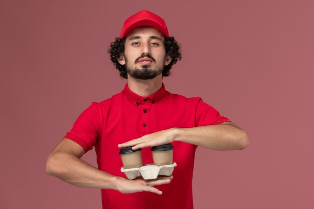 Vista frontal repartidor de mensajería masculino en camisa roja y capa sosteniendo tazas de café de entrega marrón en la pared rosa claro trabajo de empleado de entrega de servicio