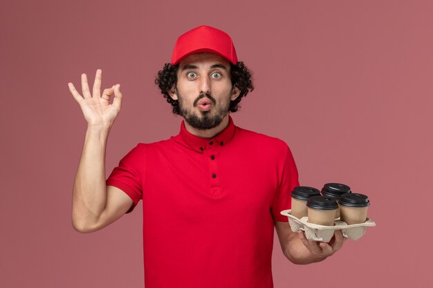 Vista frontal repartidor de mensajería masculino en camisa roja y capa sosteniendo tazas de café de entrega marrón en la pared rosa claro trabajo de empresa de empleado de servicio de entrega