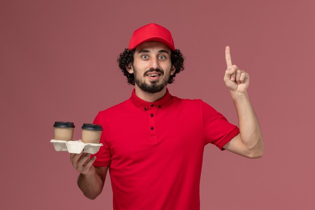 Vista frontal repartidor de mensajería masculino en camisa roja y capa sosteniendo tazas de café de entrega marrón en la pared rosa trabajador empleado de entrega de servicio