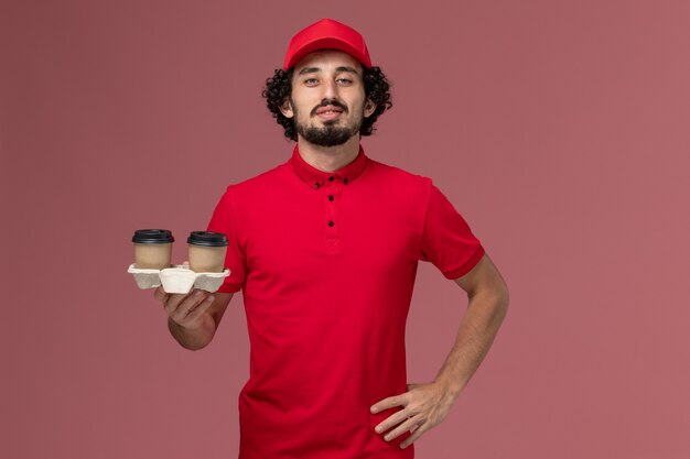 Vista frontal repartidor de mensajería masculino en camisa roja y capa sosteniendo tazas de café de entrega marrón en la pared rosada trabajo de empleado de entrega de servicio