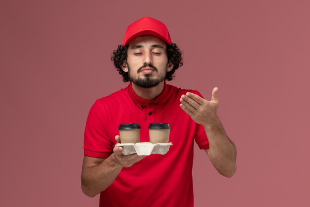 Vista frontal repartidor de mensajería masculino en camisa roja y capa sosteniendo tazas de café de entrega marrón que huelen a empleado de entrega de servicio de pared rosa