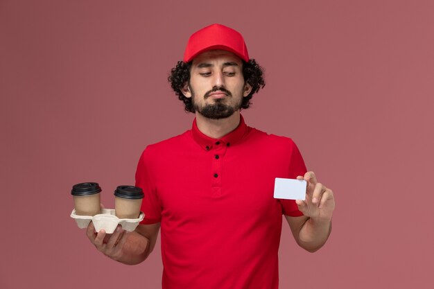 Vista frontal repartidor de mensajería masculino en camisa roja y capa sosteniendo tazas de café de entrega marrón y tarjeta en la pared rosa claro empleado de servicio de entrega masculino