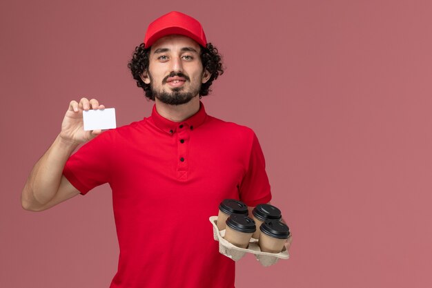 Vista frontal repartidor de mensajería masculino en camisa roja y capa sosteniendo tazas de café de entrega marrón con tarjeta de plástico en la pared rosa empleado de servicio de entrega