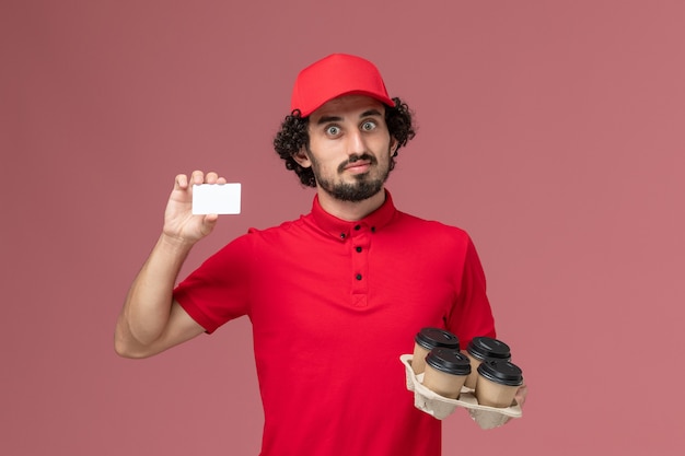 Vista frontal repartidor de mensajería masculino en camisa roja y capa sosteniendo tazas de café de entrega marrón con tarjeta de plástico en el trabajo de empleado de entrega de servicio de pared rosa