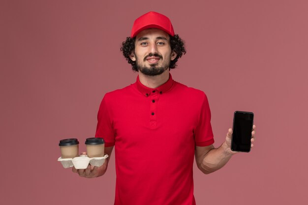 Vista frontal repartidor de mensajería masculino en camisa roja y capa sosteniendo tazas de café de entrega marrón y teléfono en la pared rosa claro empleado de servicio de entrega