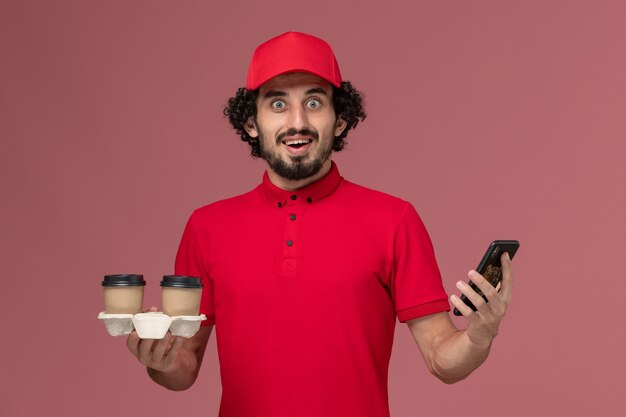 Vista frontal repartidor de mensajería masculino con camisa roja y capa sosteniendo tazas de café de entrega marrón y teléfono en la pared rosa claro trabajo de trabajador de servicio de entrega