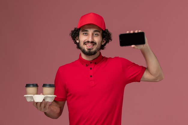 Vista frontal repartidor de mensajería masculino con camisa roja y capa sosteniendo tazas de café de entrega marrón y teléfono en el trabajo de empleado de entrega de servicio de pared rosa claro