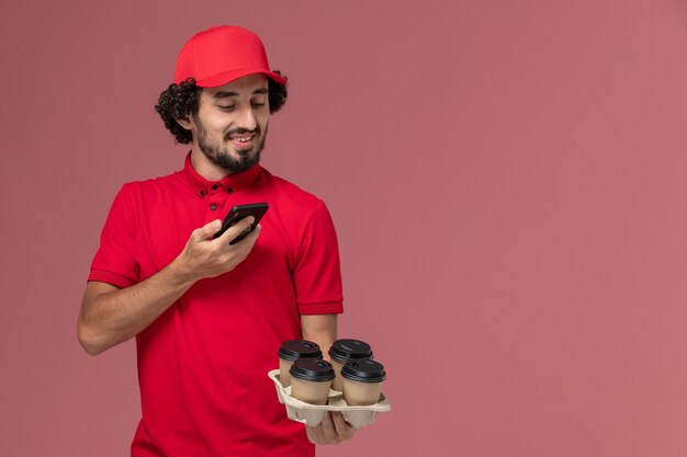 Vista frontal repartidor de mensajería masculino en camisa roja y capa sosteniendo tazas de café de entrega marrón y tomando una foto en la pared rosa claro empleado de servicio de entrega