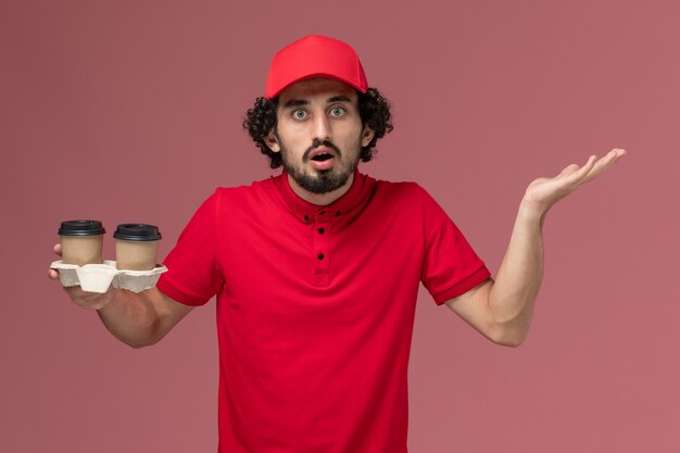 Vista frontal repartidor de mensajería masculino en camisa roja y capa sosteniendo tazas de café de entrega marrón en el trabajo de empleado de entrega de servicio de pared rosa claro