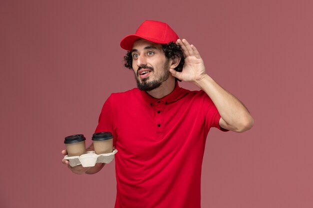 Vista frontal del repartidor de mensajería masculino en camisa roja y capa sosteniendo tazas de café de entrega marrón tratando de escuchar a un empleado de entrega de servicio de pared rosa claro