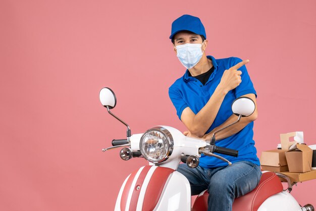 Vista frontal del repartidor preguntándose en máscara médica con sombrero sentado en scooter sobre fondo de melocotón pastel