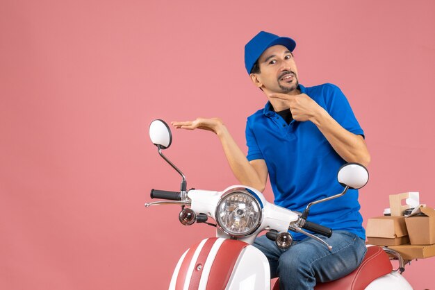 Vista frontal del repartidor sonriente con sombrero sentado en scooter y apuntando algo en el lado derecho sobre fondo melocotón pastel