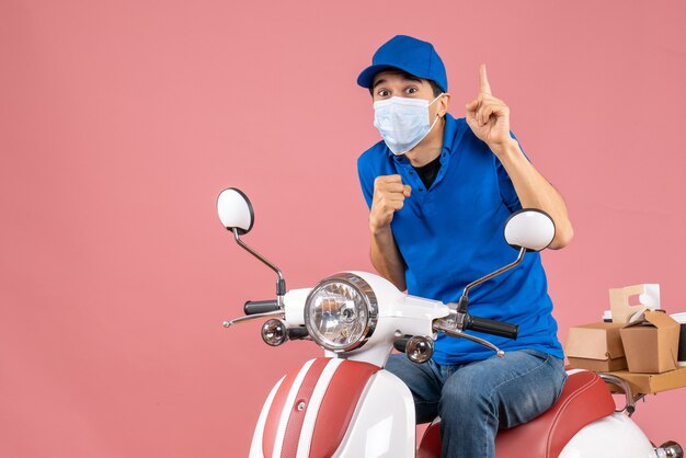 Vista frontal del repartidor sorprendido en máscara médica con sombrero sentado en scooter y apuntando hacia arriba sobre fondo de melocotón pastel