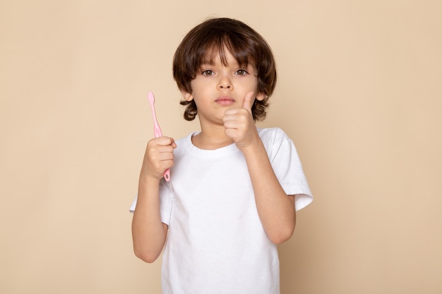 Vista frontal del retrato, niño niño con cepillo de dientes en sus manos en camiseta blanca en escritorio rosa