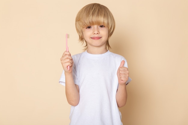 Vista frontal del retrato, sonriente niño rubio lindo adorable con cepillo de dientes en camiseta blanca y pared rosa