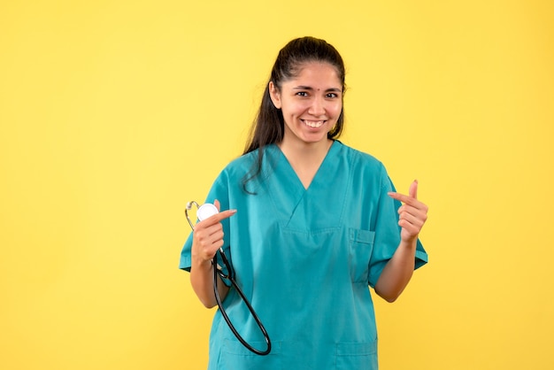 Vista frontal riendo doctora sosteniendo estetoscopio en su mano sobre fondo amarillo