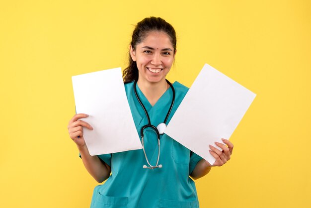 Foto gratuita vista frontal riendo mujer médico en uniforme sosteniendo papeles con ambas manos sobre fondo amarillo