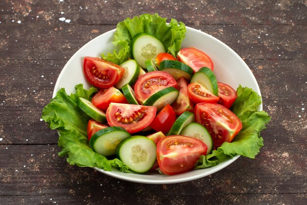 Vista frontal rodajas de tomates con pepinos dentro de un plato blanco con ensalada verde en marrón, comida vegetal fresca comida sald