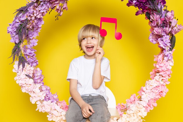 Una vista frontal rubia sonriente niño en camiseta blanca con nota rosa en la flor hecha soporte en el escritorio amarillo