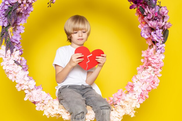 Una vista frontal rubio lindo chico en camiseta blanca con forma de corazón sentado en la flor hecha soporte en el piso amarillo