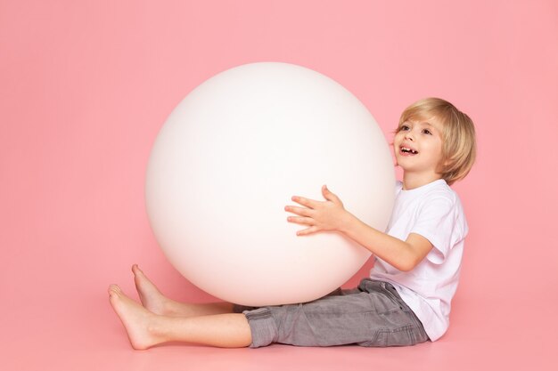 Una vista frontal rubio niño feliz en camiseta blanca jugando con pelota blanca en el escritorio de color rosa