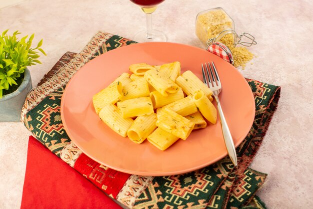 Una vista frontal de la sabrosa comida de pasta italiana dentro de la placa rosa junto con la pasta cruda y una copa de vino en la alfombra de colores y rosa